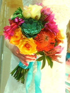 Bright golden and orange roses accompany succulents, orange proteas, and coral dahlias in this bridal bouquet. 